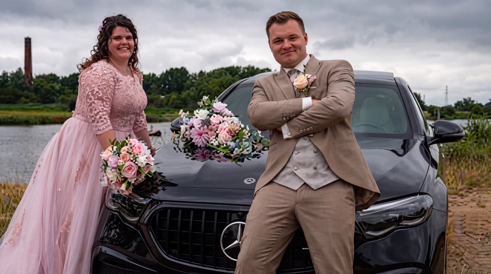 Bruidspaar leunt tegen de trouwauto met hun bruidsboeketten